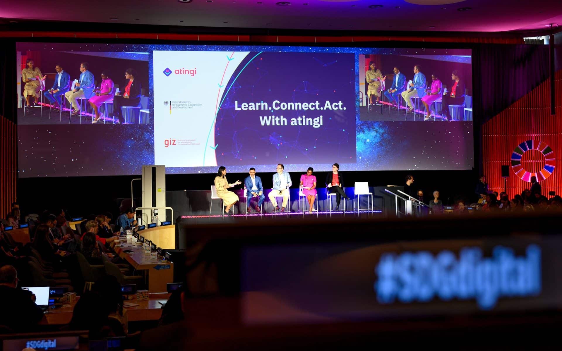 Conference room with speakes on stage with atingi information projected on screens.